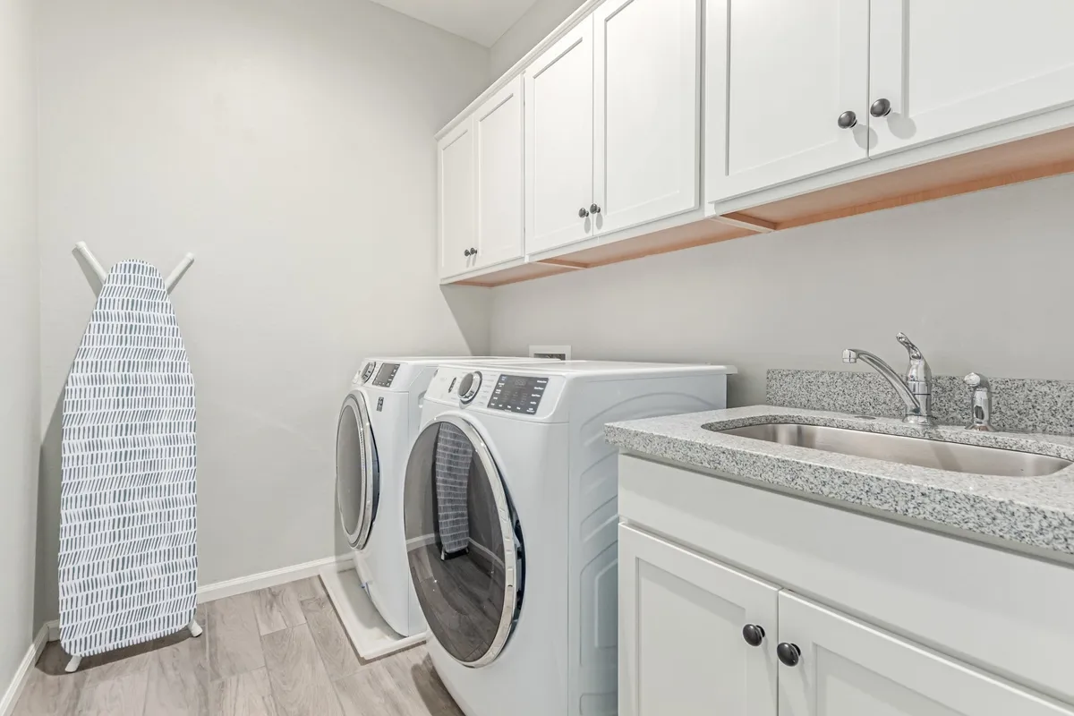 A bright and functional laundry area equipped with modern washing machines, a large sink, and an ironing board. The space is organized for convenience and ease of use, suggesting the comfort of in-home laundry facilities.