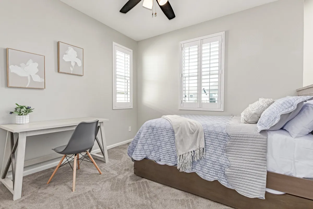 A serene bedroom featuring a comfortable bed, a dedicated desk workspace, and framed art. The room catches natural morning light, offering a calm setting for both work and relaxation.