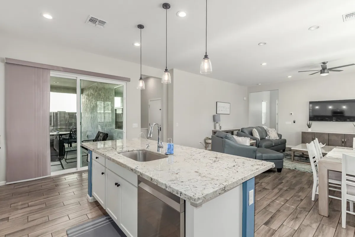 A modern, open-concept area with a stylish kitchen island in the foreground and a comfortable living room seating area beyond. Pendant lights and a large sliding glass door add brightness and a sense of openness, seamlessly blending spaces for cooking, dining, and relaxing.