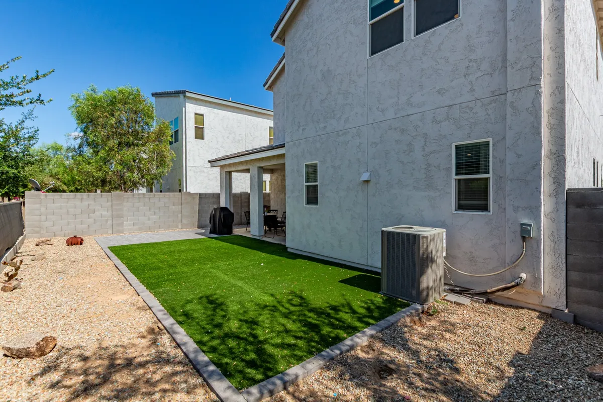A sunny exterior view showcasing an artificial grass lawn, a welcoming patio area with a grill, and the home's exterior walls. The space is set up for outdoor dining, barbecuing, and relaxed gatherings, perfect for groups of friends spending time together outside.