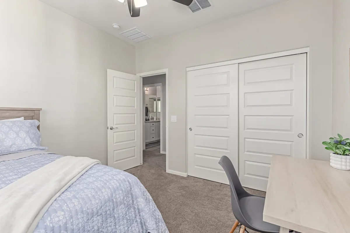 A calm bedroom with blue floral bedding is shown, complete with a dedicated desk and chair for work, and an open door leading to an en suite bathroom. Natural light fills the space, emphasizing its functionality for both rest and productivity.